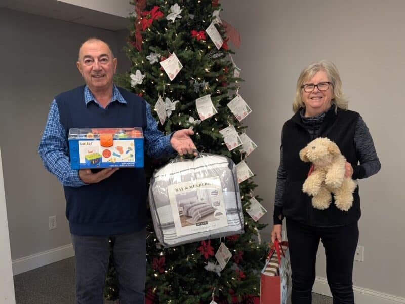 Two people holding gifts stand in front of the Giving Tree.