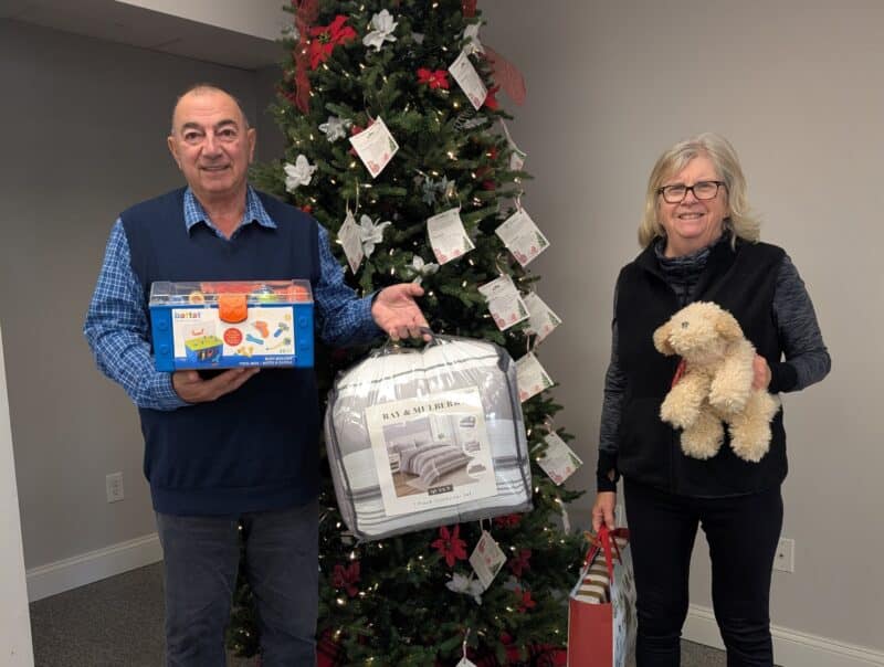 Two people holding gifts stand in front of the Giving Tree.